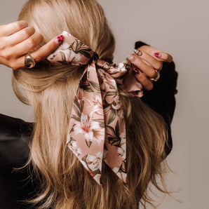 A person with long blonde hair is shown from behind, wearing a silk twilly scarf with a floral pattern tied in their hair.